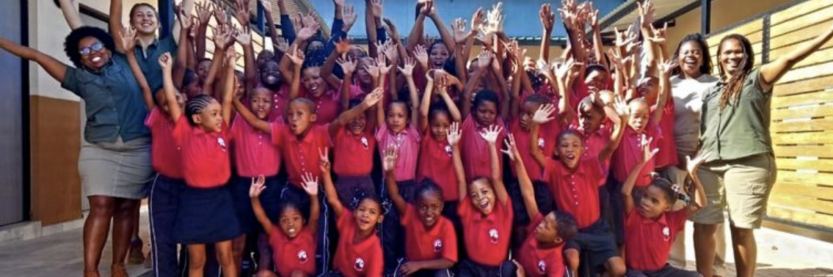 Gruppe af San-børn og lærere, der løfter armene og smiler i skolegården på deres folkeskole i Namibia – et symbol på glæde og fælles succes.