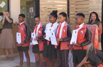 Fem San-elever i skoleuniform modtager deres certifikater ved en afslutningsceremoni på en lokalskole i Namibia, omgivet af deres lærere.