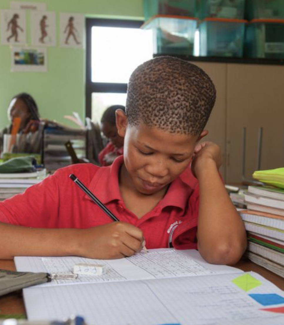San-elev fordybet i en skriveøvelse i et klasselokale i Namibia, omgivet af hæfter og skolematerialer.