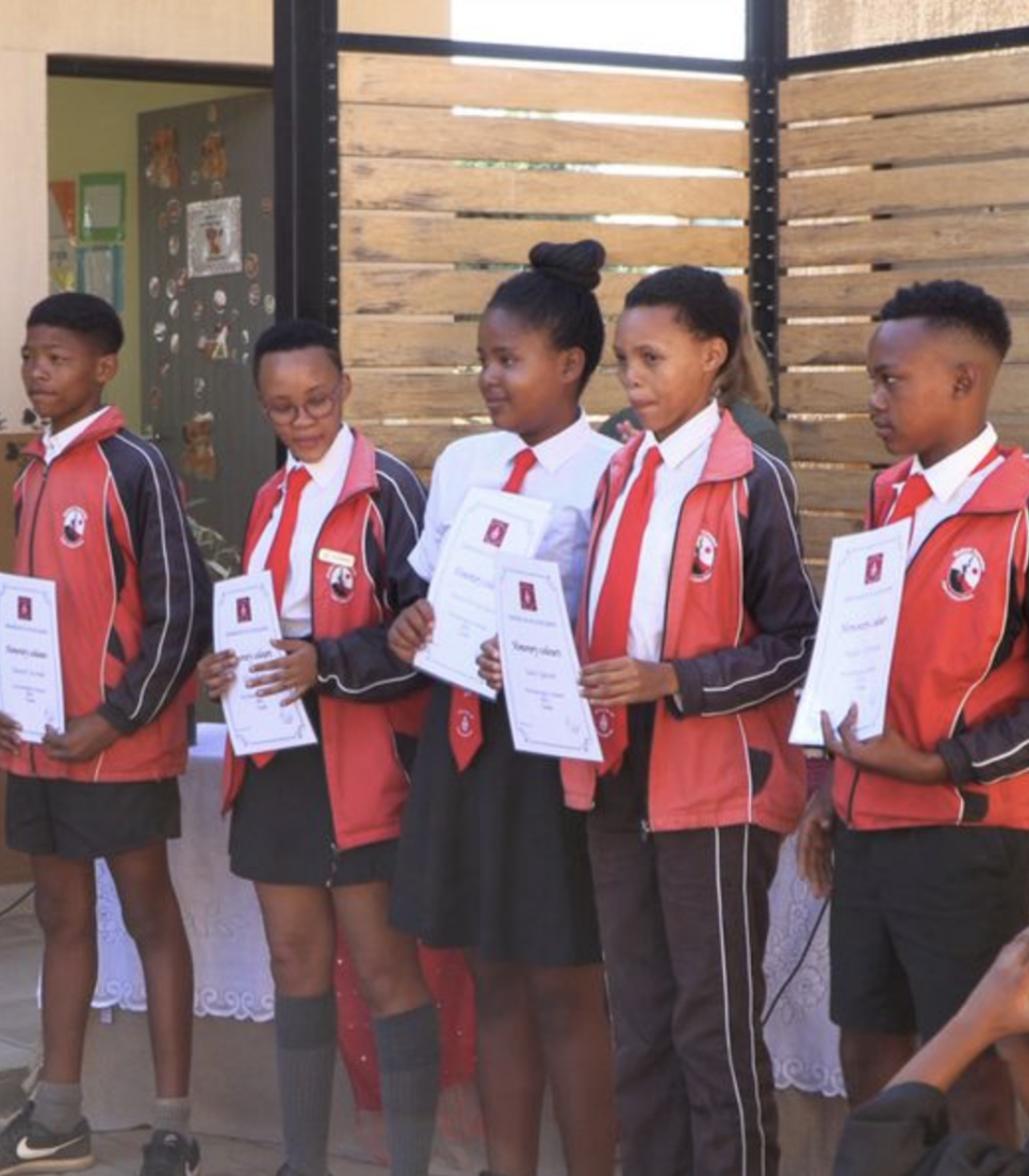 Fem San-elever i skoleuniform modtager deres certifikater ved en afslutningsceremoni på en lokalskole i Namibia, omgivet af deres lærere.
